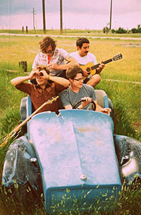 four men sitting in junked remains of antique car body, one playing a guitar