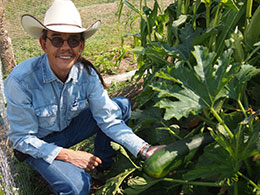 man in cowboy hat squatting beside plant, holding large zucchini