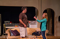 woman showing something to a man in front of a sofa on a stage set