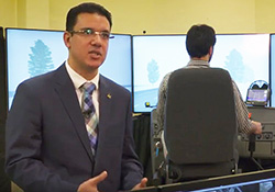 man standing in front of video screens with another man seated at the screens