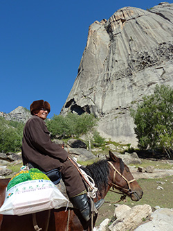 person on horse with something slung behind saddle and rocky outcropping in background