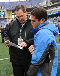 two men looking at a clipboard on the sidelines of a football game