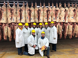 people in white coats and hard hats with rows of meat hanging behind them