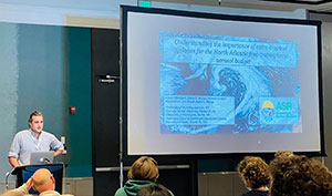 man speaking to an audience with a large screen in the background