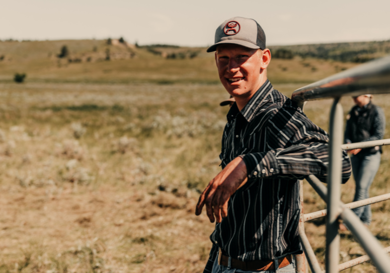 Young man leaning on ranch gate wearing a baseball cap and striped shirt. Wyoming prairie bedhing him. 