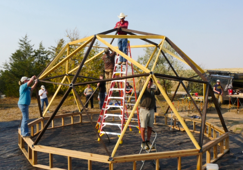 Educators use ladders to assemble the wooden frame of a geodesic dome outdoors