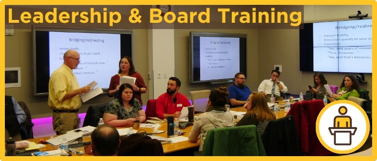 a group of people around a conference table listen to 2 people standing and explaining