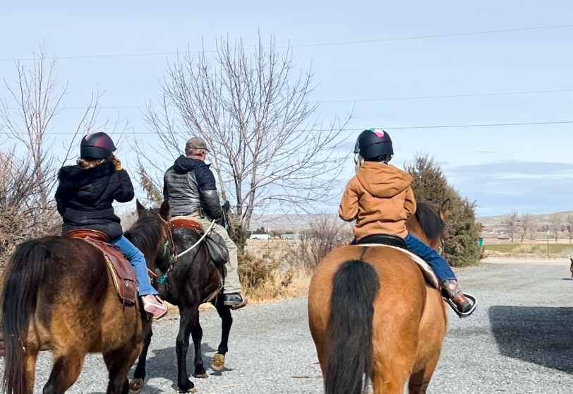 Three youth wearing helmets ride horses outdoors along a rural trail.