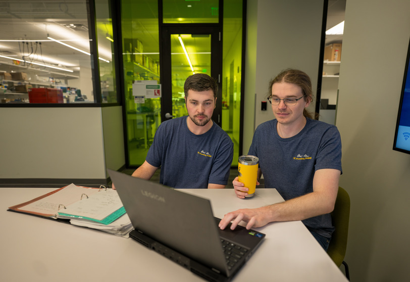 two students sit at a table and look at a laptop