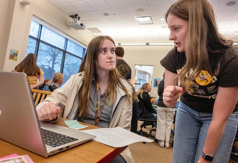 A student sits at a table at a laptop while another person is speaking to them