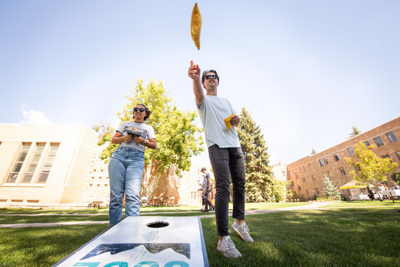 A sunny outdoor scene at a campus with two young adults playing cornhole; a man throws a yellow bean bag, while a woman watches nearby, both dressed in casual summer clothes.