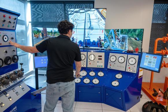 A person operates a blue control panel with gauges and screens in a simulation room, displaying images of machinery operations.