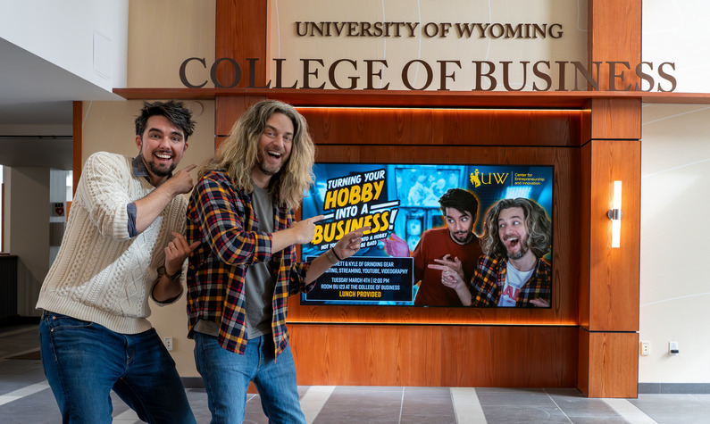 Two men in the College of Business point toward a video screen