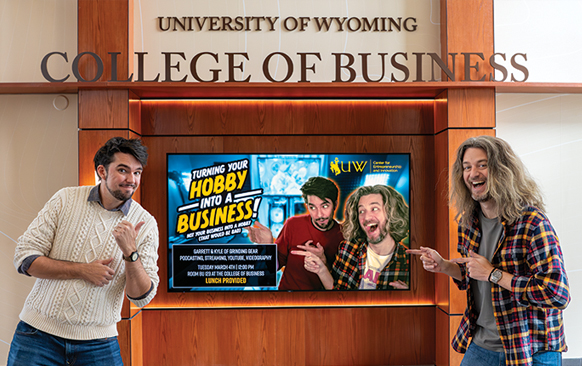 Two man pointing at a screen advertising their speech in the UW College of Business