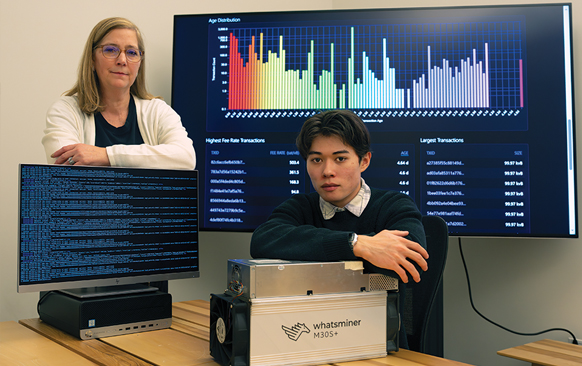 Staff member and a student pose with multiple screens and a bitcoin miner.