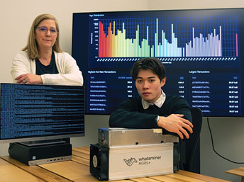 Staff member and a student pose with multiple screens and a bitcoin miner.