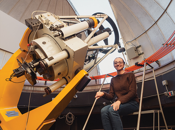 female student sitting by a large telescope