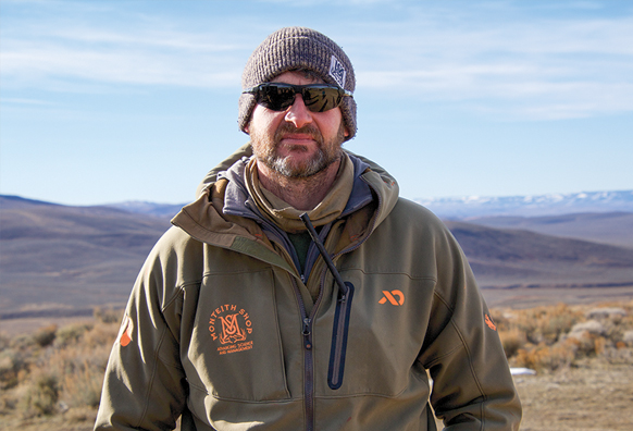 A man wearing a brown jacket, hat, and sunglasses poses outside