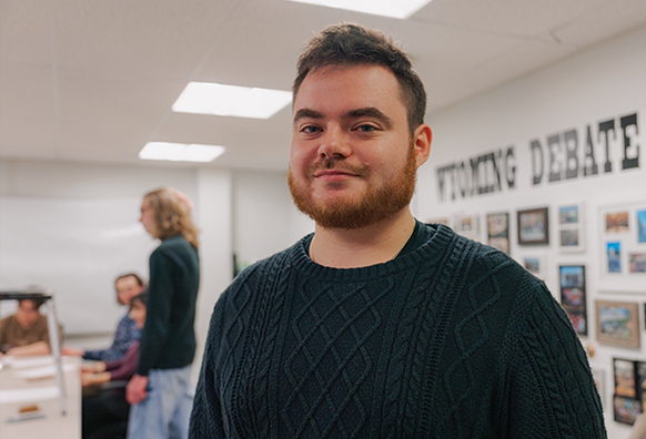man stands in debate room with students in the background