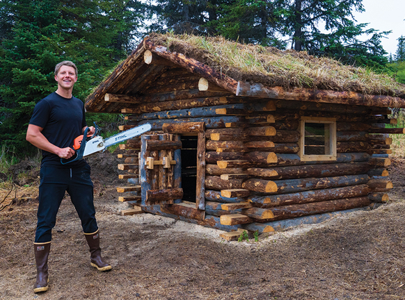 Person with a chainsaw near a log cabin