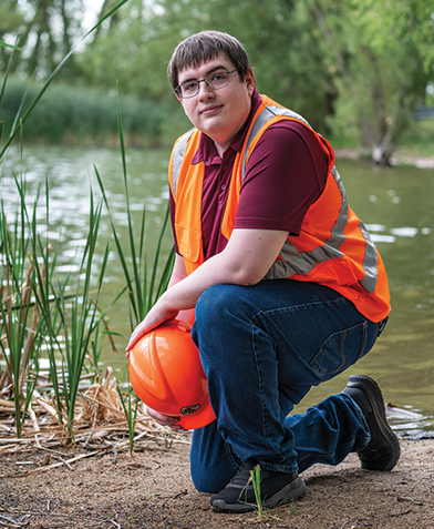 Person in an orange safety vest holding a hardhat by a lake