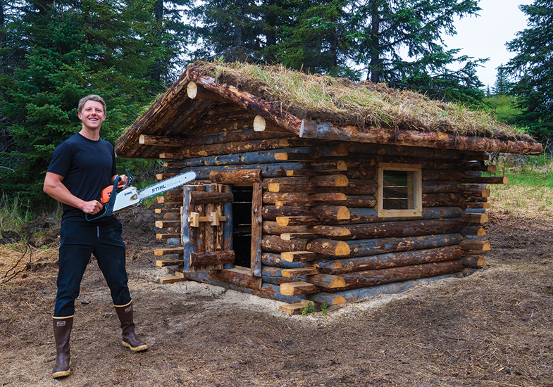 person holding a chainsaw by a log cabin
