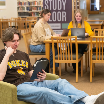 a group of people in a library using different types of computer devices such as laptops and tablets.