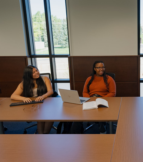 Two students sit at a table discussing topic with their computers in front of them.