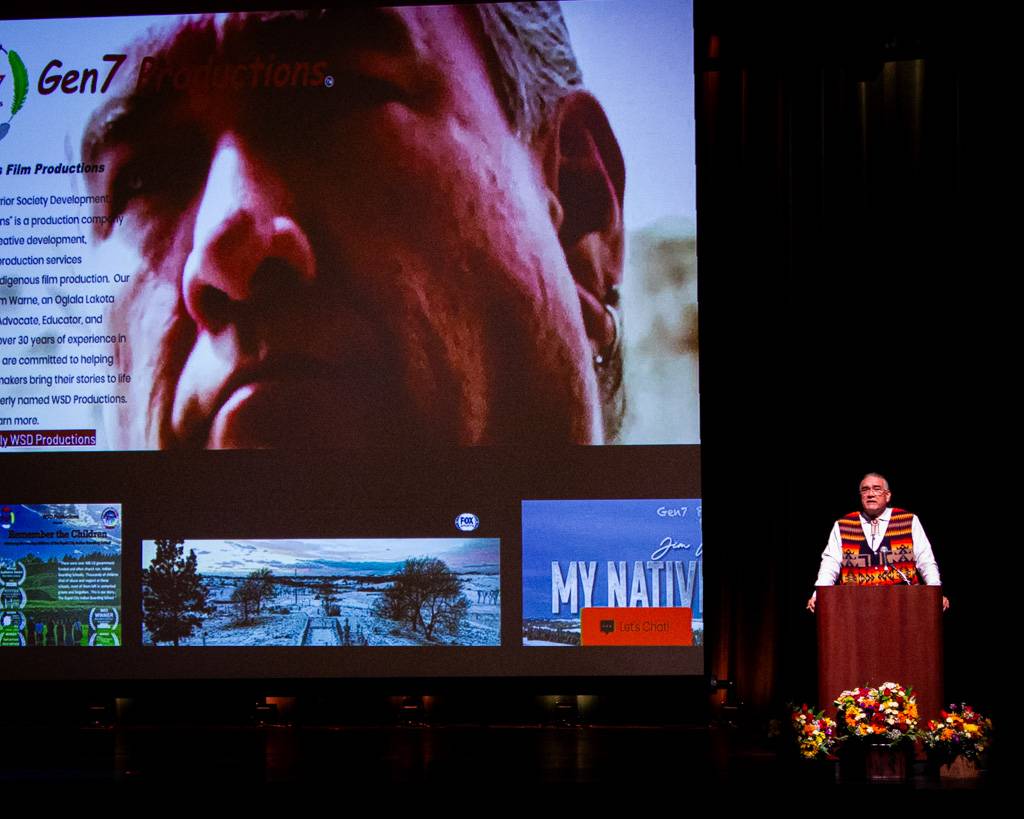 Jim Warne standing behind a podium with his award-winning-film, 7th Generation, playing behind him.