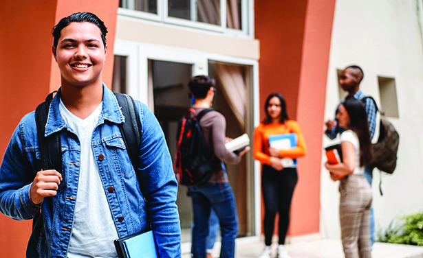 Student smiles for the camera. In the background is a school enterence with 4 other students conversing