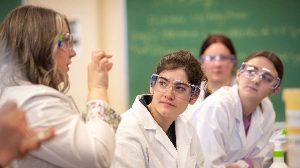 Instructor explaining concept with hands to three students in a laboratory in lab coats and goggles