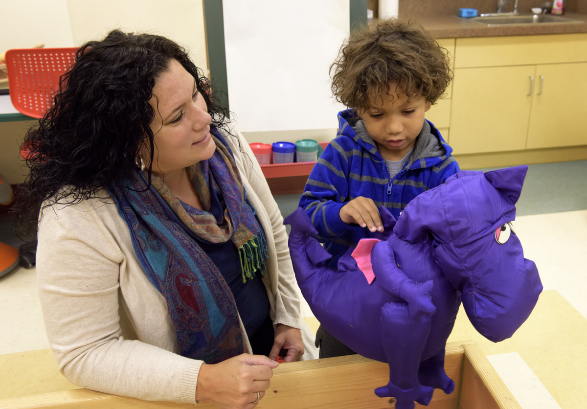 Adult female teacher speaking with small boy and there is purple dinosaur stuffed animal