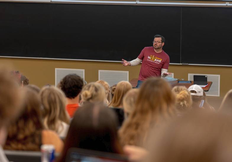 A man wearing a red WWAMI shirt speaking in-front of a large crowd.
