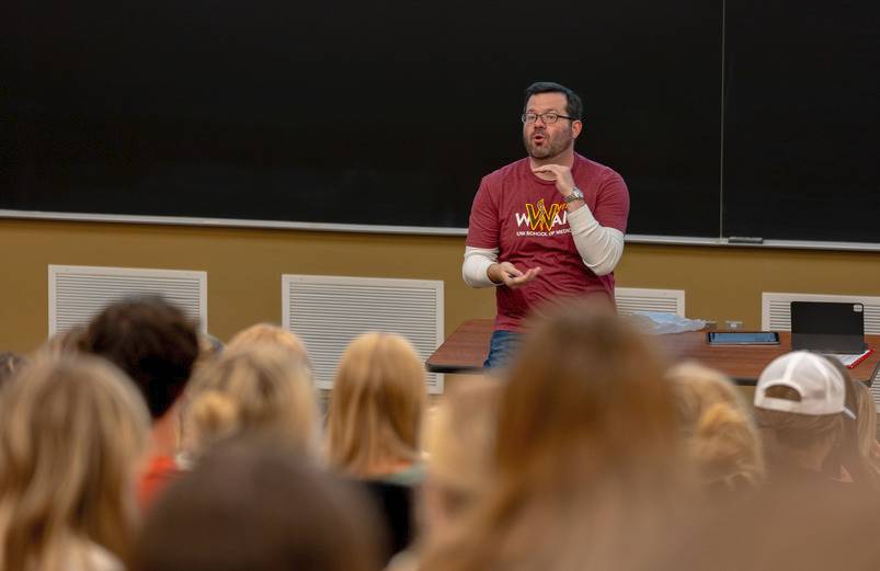 A man wearing a WWAMI t-shirt giving a lecture in front of a large group of students.