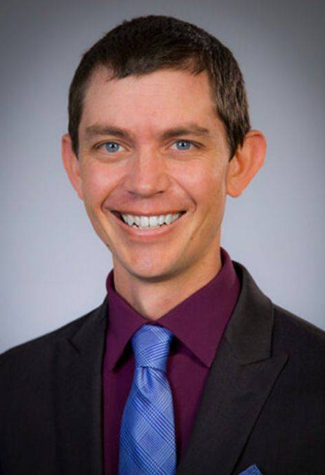 Aaron Spurlock, wearing a black suit jacket, a dark purple button-up shirt, and a blue tie.