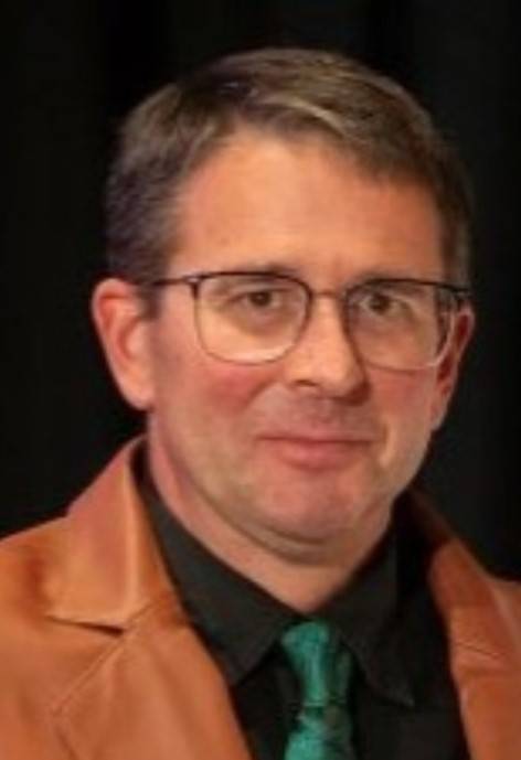 Cam Walker, wearing a tan leather jacket, a black button-up shirt, and a green tie.
