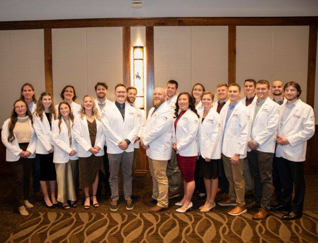 A group of 20 students wearing white doctor coats, standing together and smiling at the camera.