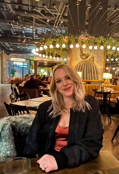 Shayna Bauer, wearing a black jacket and a dark peach blouse. Sitting in a brightly lit resturant.