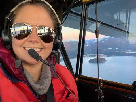 Jenni Ebersberger, wearing a red snow jacket and sunglasses in the cockpit of a small plane.
