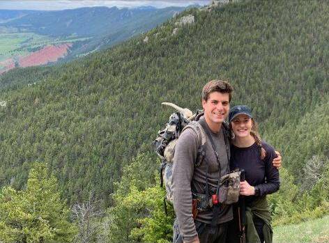 Andrew with his wife, hiking in the Big Horn Mountains. They are standing on a hill with a pine forest in the distance.