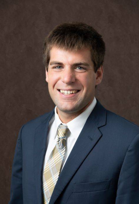 Andy Quinn wearing a dark blue suit jacket, white button-up, and metallic gold tie.