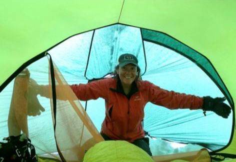 Heidi wearing a bright orange snow jacket, sitting in a green and blue tent.