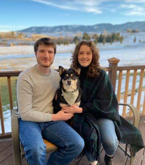 Andrew sitting with his wife on a wooden deck, they are holding a small tri-colored dog between them.