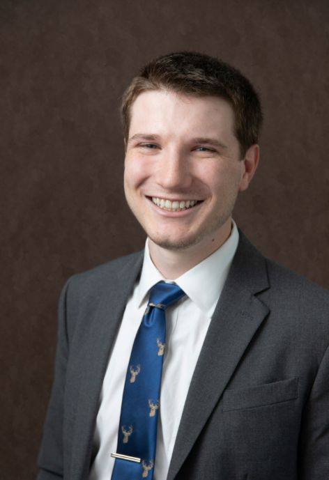 Andy White wearing a grey suit jacket, white button-up, and a blue tie.