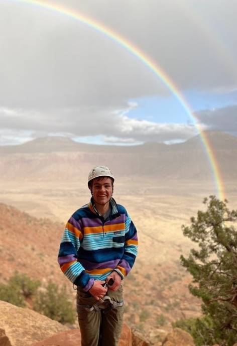 Brad wearing a rockclimbing helmet, standing in a red desert and there is a rainbow in the background.