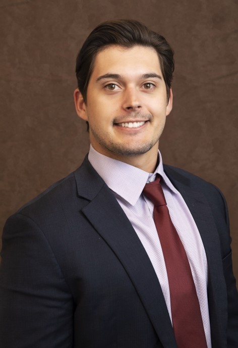 Carson Asher wearing a dark blue suit jacket, lavendar button-up, and wine colored tie.