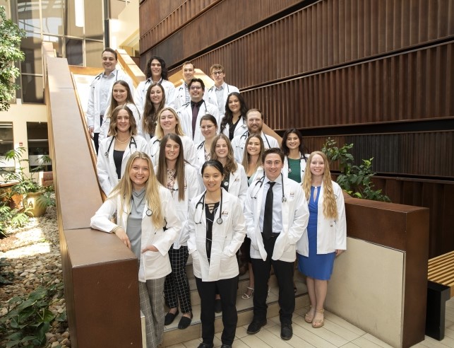 A group of 20 students wearing doctor coats and standing together on a staircase.