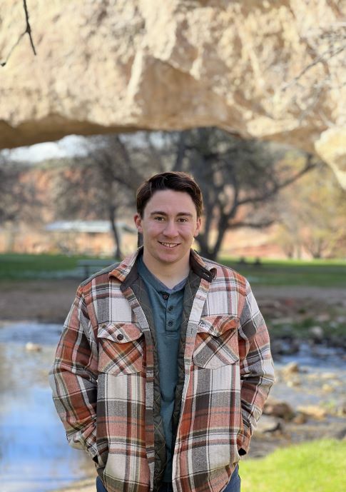 Noah standing outside wearing an orange plaid jacket. There is a stone arch and small pond in the background.