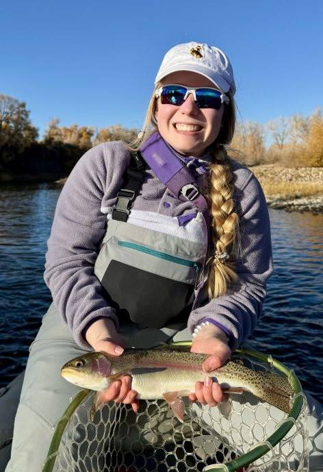Cassidy wearing fishing gear, standing in a boat, and holding a rainbow trout.