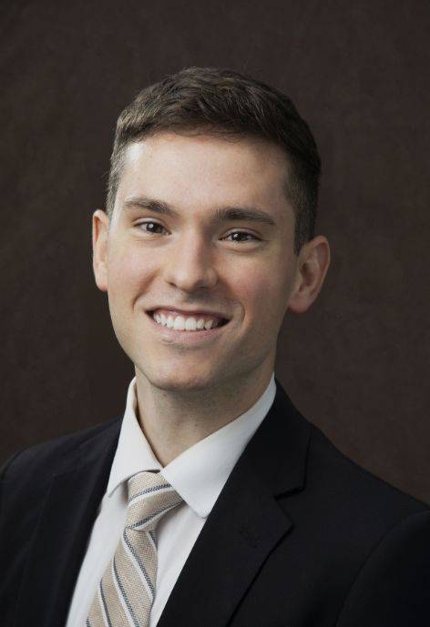 Jordan Moore wearing a black suit jacket, white button-up, and gold patterned tie.